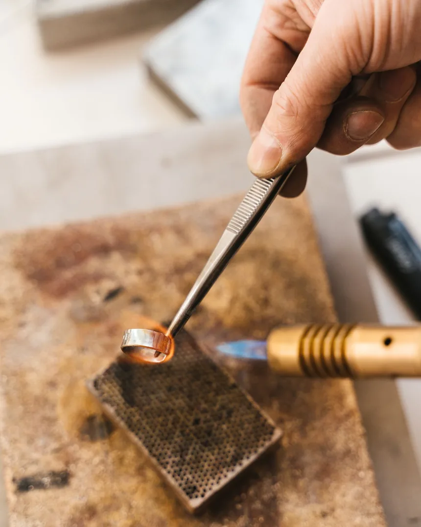 Anillo de metal sujeto con pinzas sobre placa de soldar mientras la llama del soplete calienta la unión